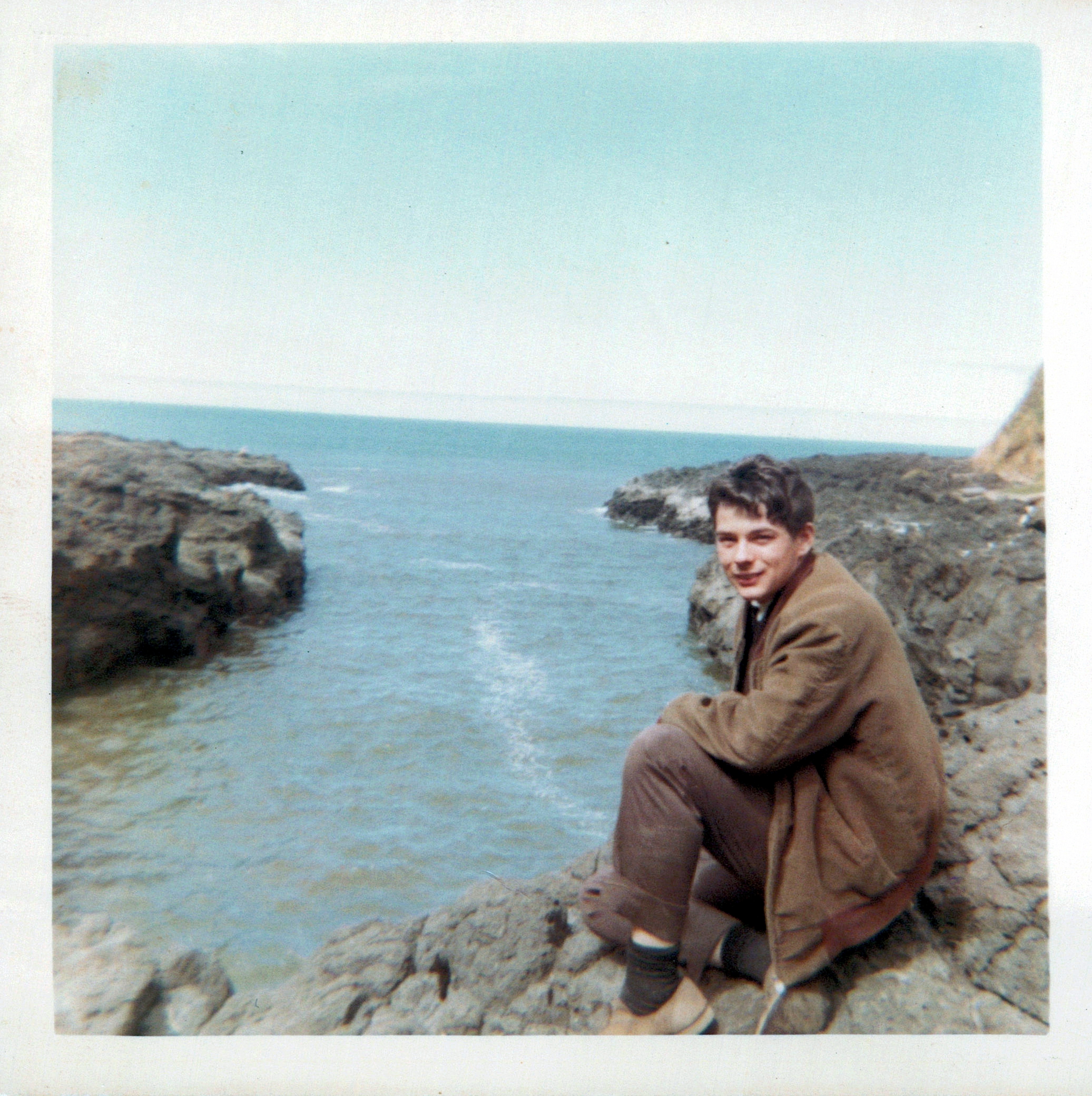 A photo of David Richard Kim Sigurdson in a brown coat, sitting on the rocky shore that borders an ocean.