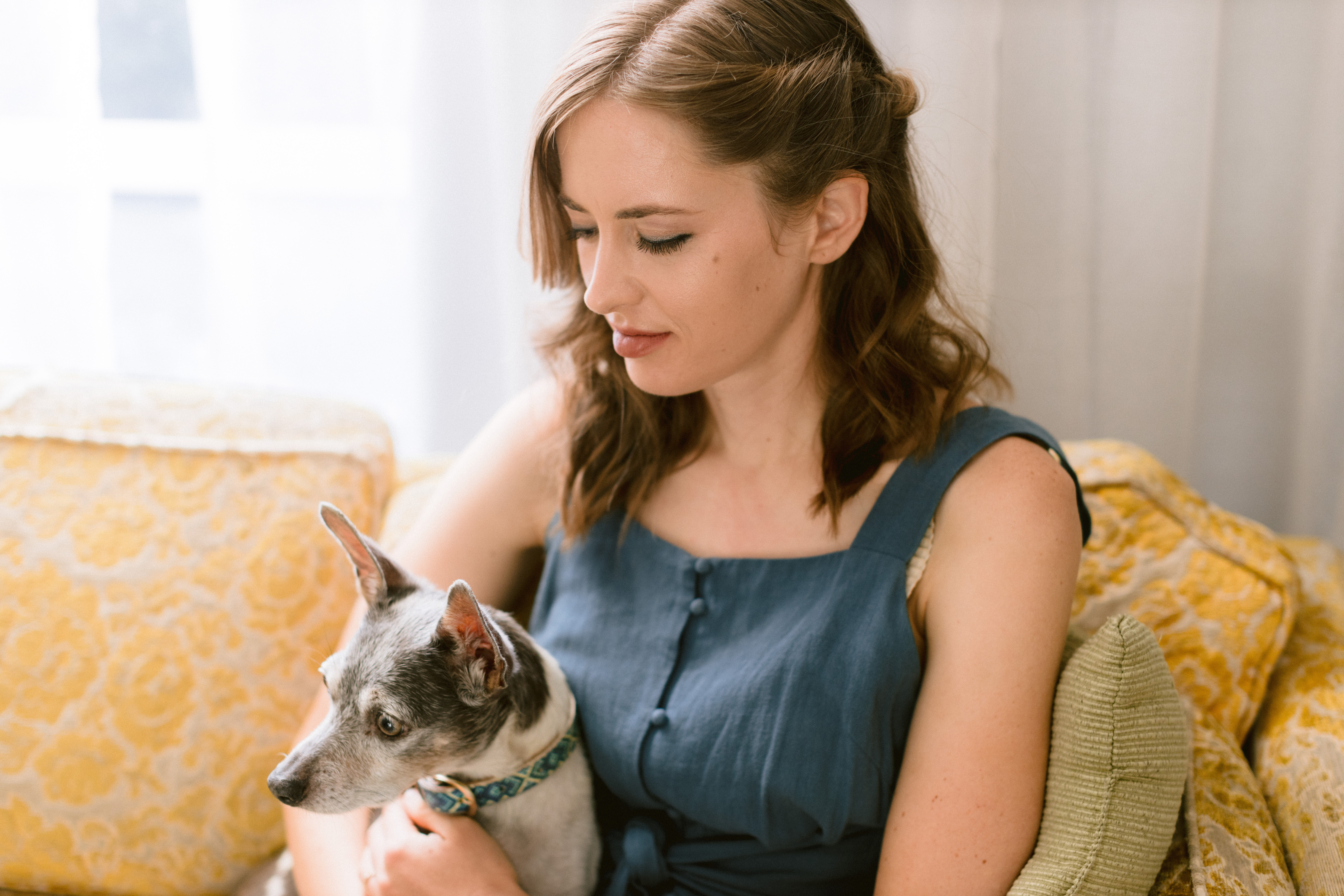 Carrie Grinstead looks down while she sits on a yellow couch, a small dog in her arms.