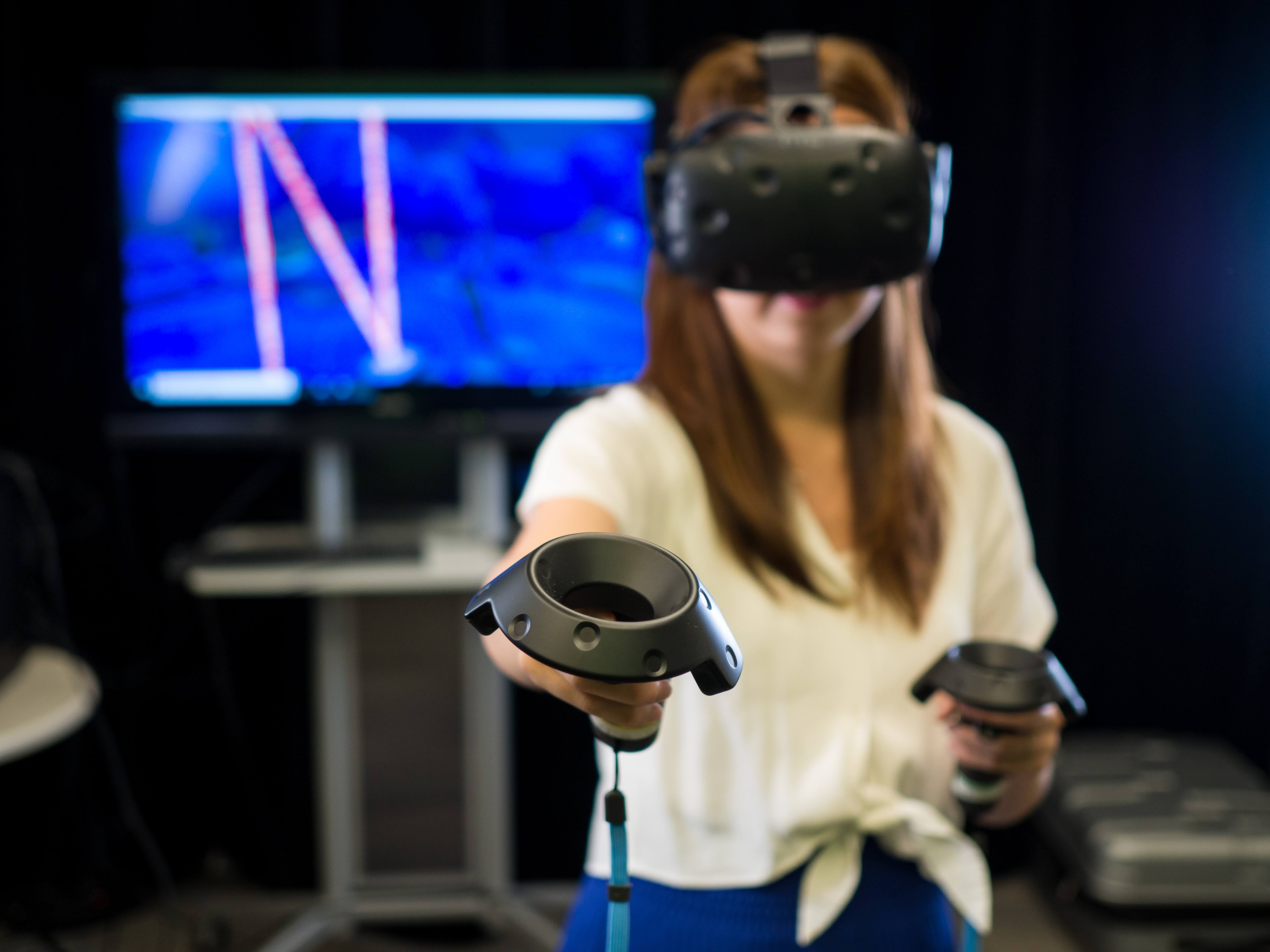 A woman with a virtual reality headset, holding virtual reality joysticks, moves forward.