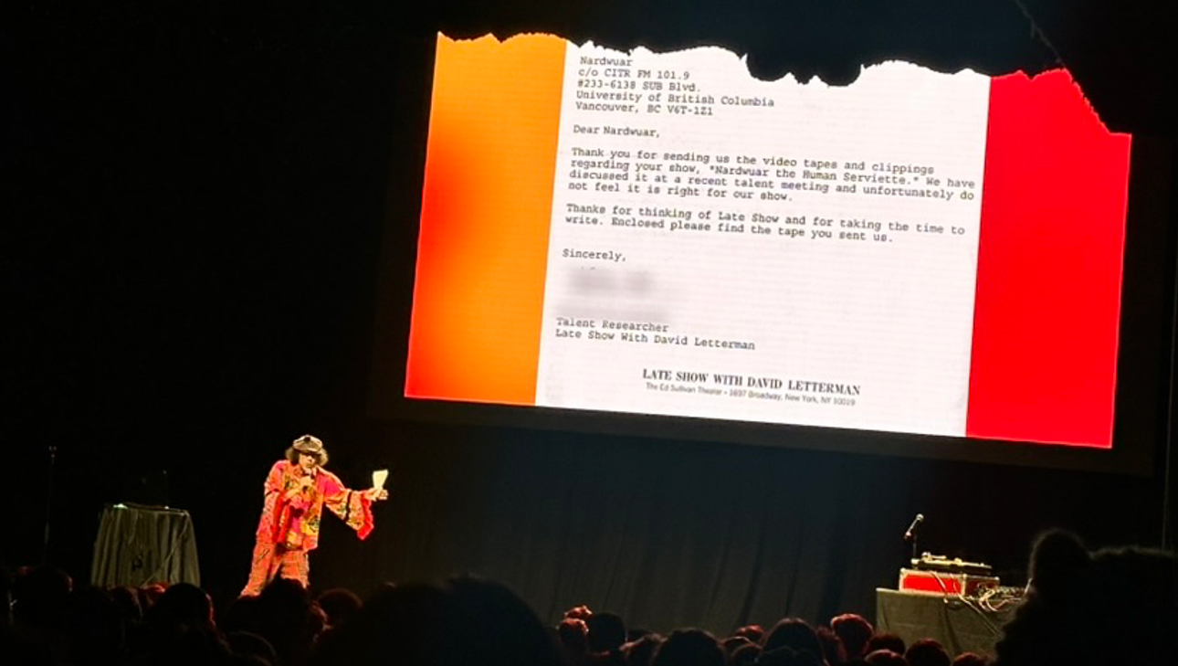 Nardwuar on the stage, with an excerpt of his rejection letter from The Late Show with David Letterman onscreen