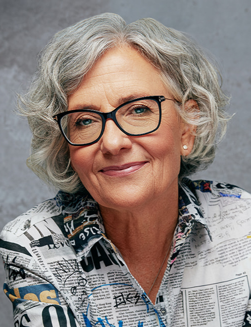 A headshot of Barb Howard with curly grey hair, black glasses, and a newsprint-patterned shirt