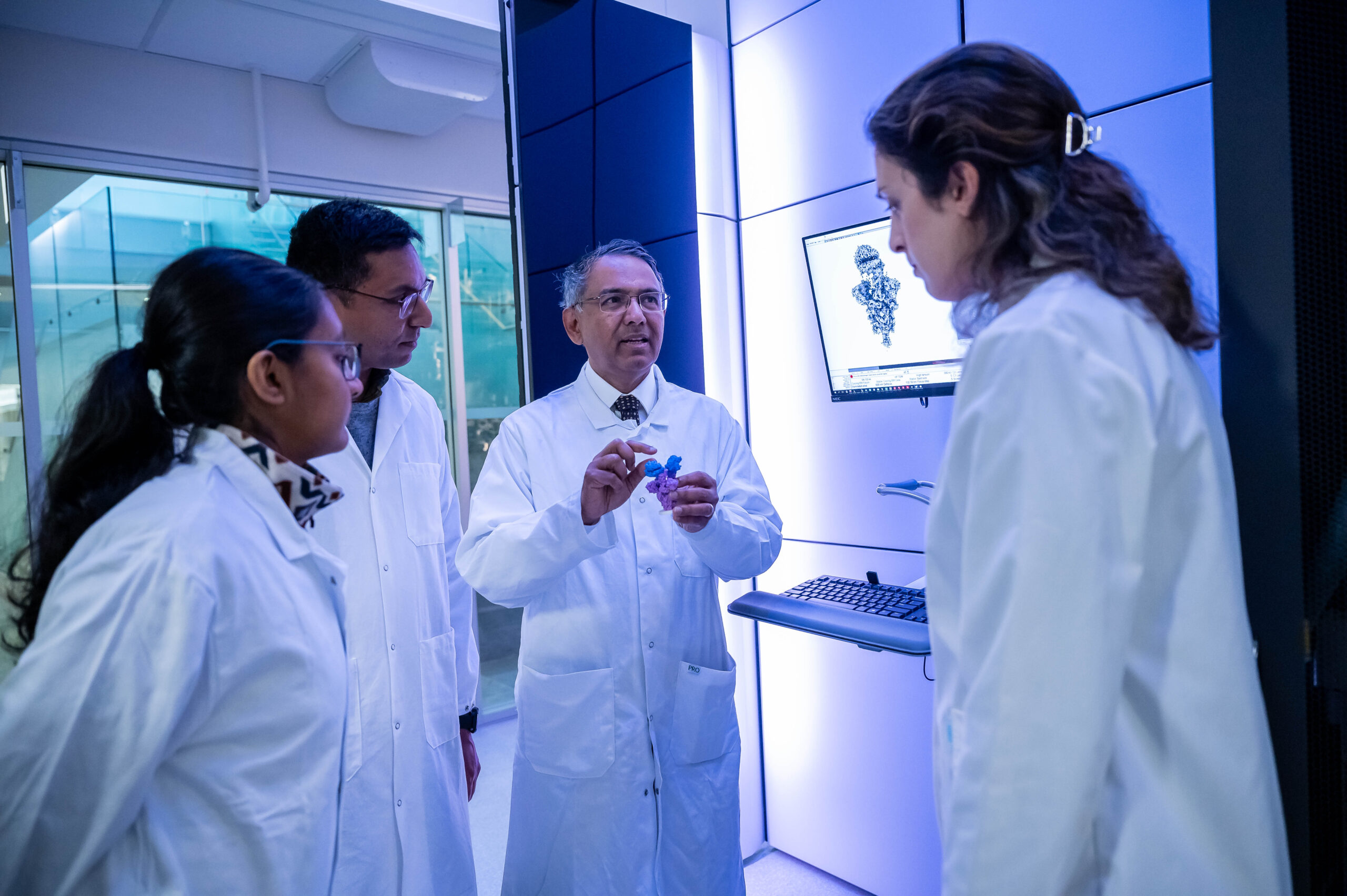 Man in lab coat speaks to three others in lab