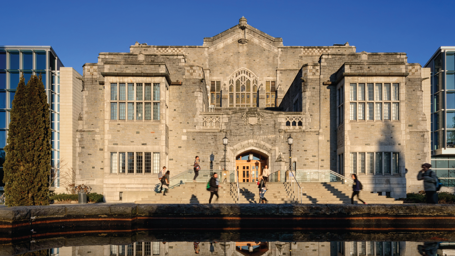 An old stone library with students passing in front of it and a fountain in the foreground