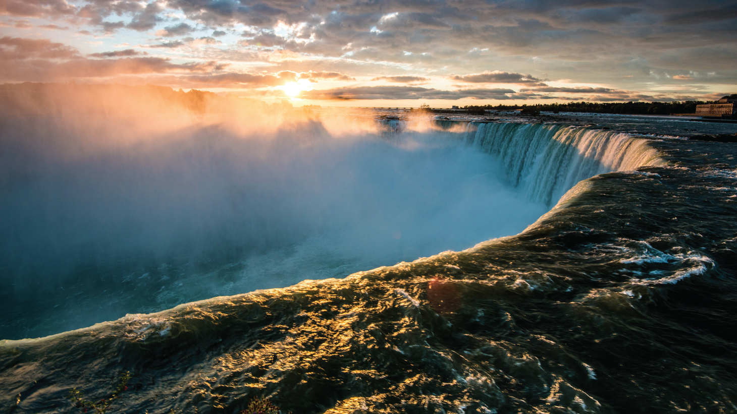 The sun rises through the mist over a large curved waterfall