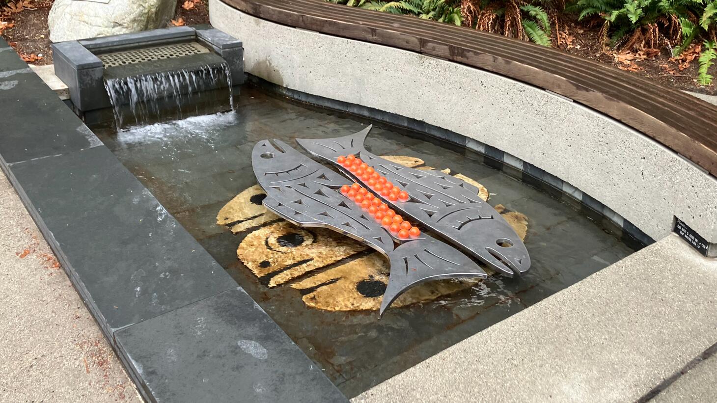 The artwork is a fountain featuring two pairs of salmon: one beneath the water, and another floating above the surface, which are made of metal and have orange glass eggs filling their bellies.