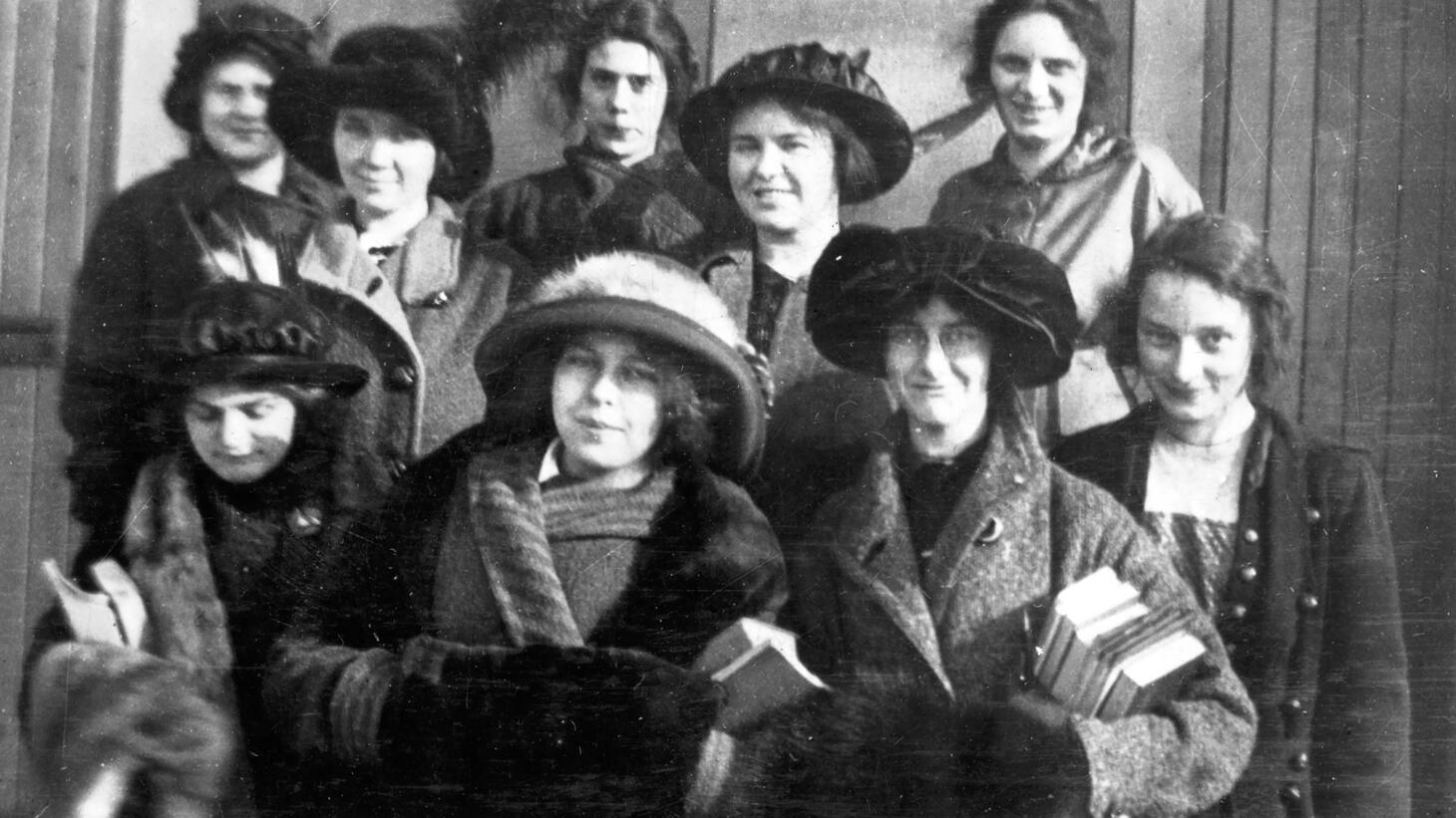 Black and white image of a group of female students wearing thick wool coats, hats decorated with fur and feathers, and carrying books