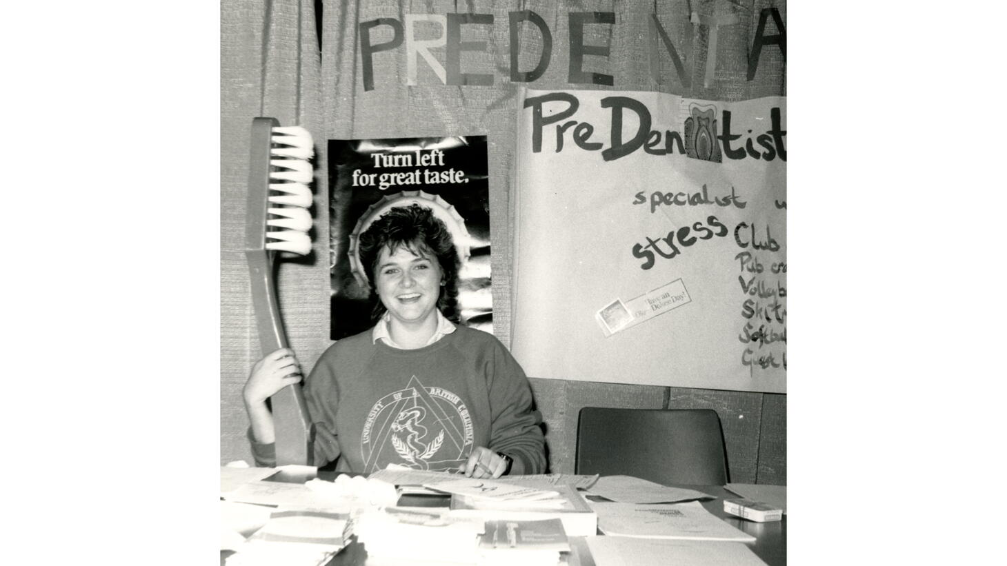 Black and white photo of a student with a mullet wearing a collared shirt under a graphic sweater holding a giant tooth brush at the Pre-Dental Society booth