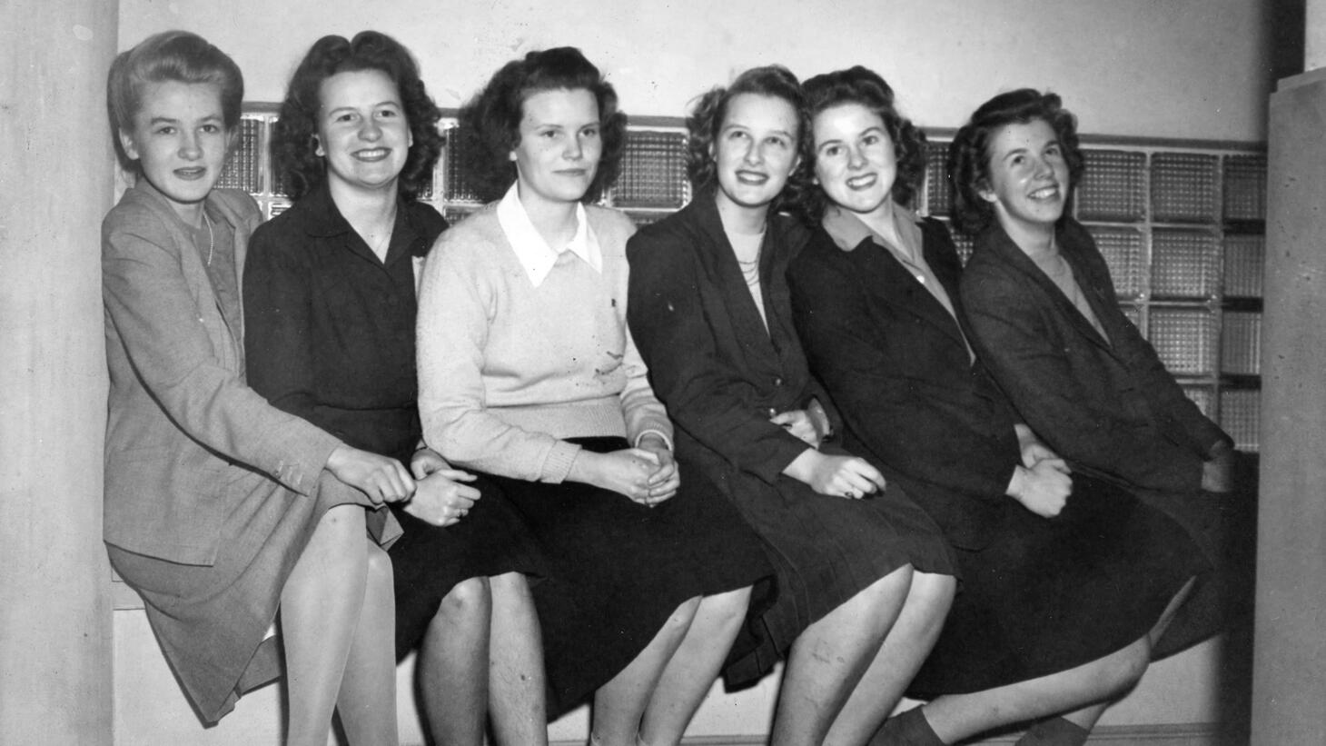 Black and white image of women with curled shoulder-length hair in knee-length skirts and suit jackets posing on a bench