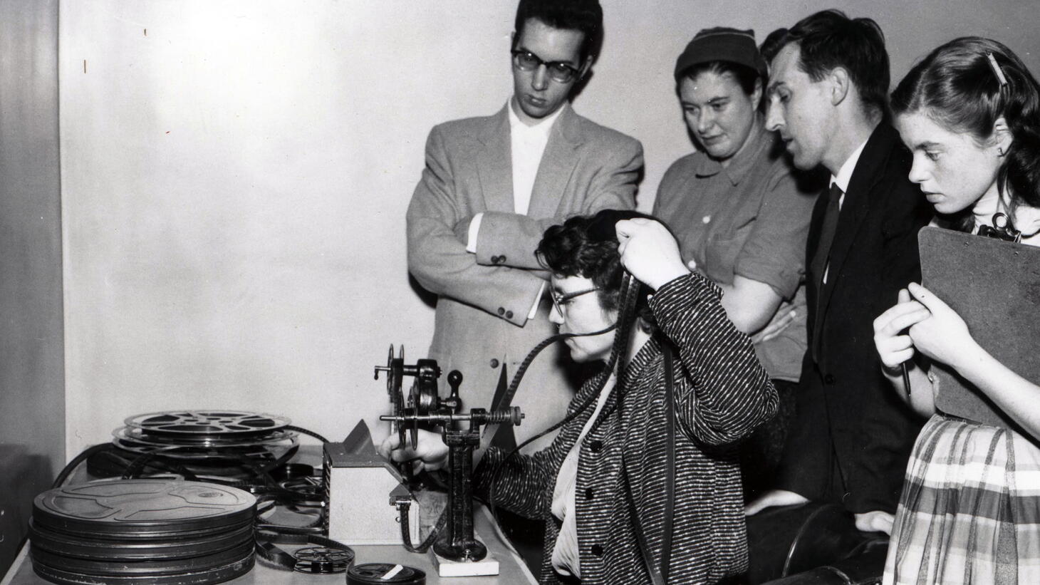 Black and white image of students watching as a teacher demonstrates the use of film, the male students are wearing suits and the female students are wearing long skirts and collared shirts