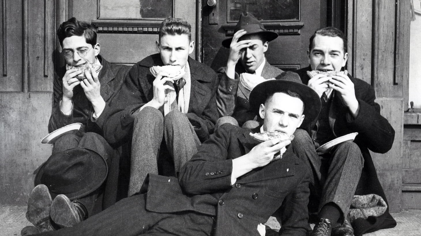 Black and white image of a group of short-haired young men in full suits and brimmed hats eating lunch on the floor
