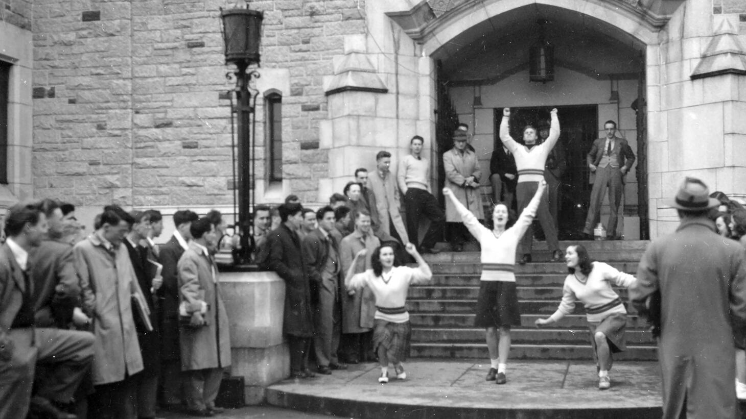 Black and white photo of students competing in an event outside Main Library, men with short hair in long coats, hats, and collared shirts, women in knee-length skirts and knit sweaters with shoulder-length curled hair