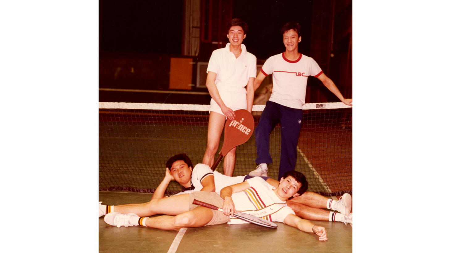 Colour photo of four male students posing in front of a tennis net in polo shirts and ring-neck tees, athletic shorts and sweatpants