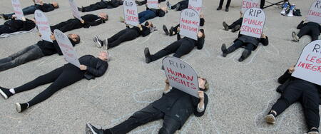 People dressed in black lying on the ground holding tombstone-shaped signs with "RIP" messages related to democracy