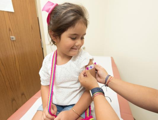 Girl sits in a clinic watching healthcare worker apply band-aid to her shoulder