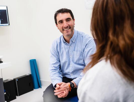Smiling doctor in room (facing a woman slightly off-camera) with monitor displaying brain scan images