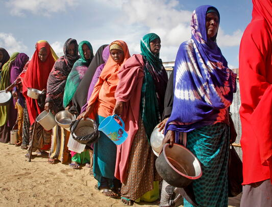 African women in shawls carrying empty containers queue up in desert 