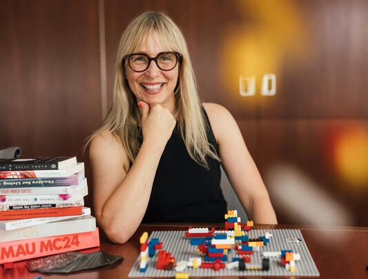 Dr. Kari Marken sits at a desk with lego, smiling at the camera.
