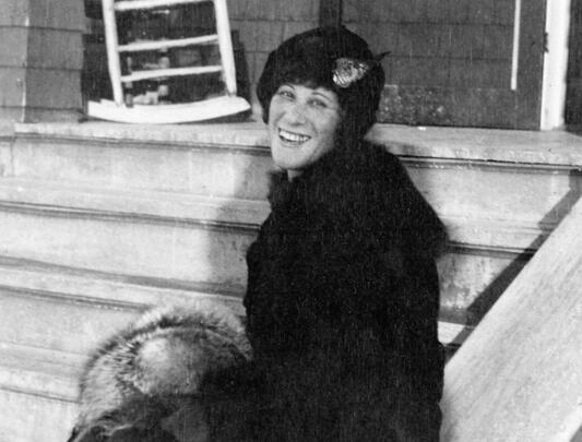 Woman in hat and coat turned sideways smiles on steps in black-and-white photo