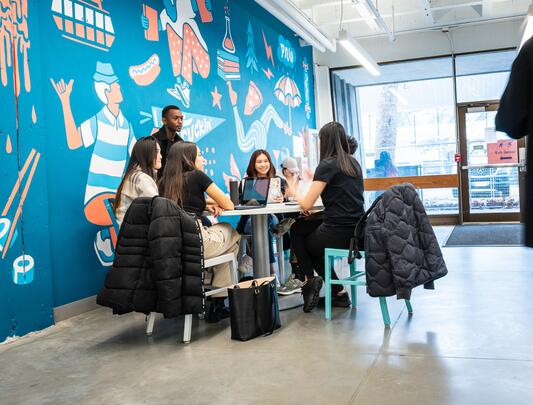 Five students sitting at a table next to wall with mural
