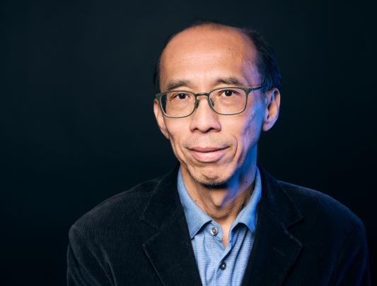 A headshot of Dr. Raymond Ng with dark green glasses, short black hair, and a blue collared shirt against a black backgroun.