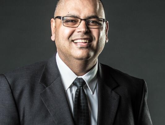 A headshot of Alex Sangha wearing black rectangular glasses and a black suit.