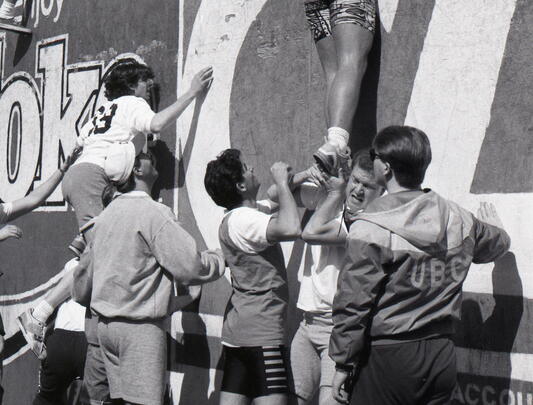 A black and white photo of students pushing each other up a wall.