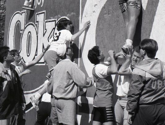 A black and white photo of students pushing each other up a wall.