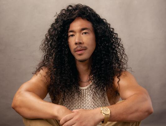 A photo of Cole Walliser with long, curly black hair and a gold watch sitting on the ground