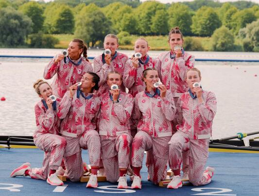 The Canadian women's rowing eight team pose on a dock while kissing their silver medals