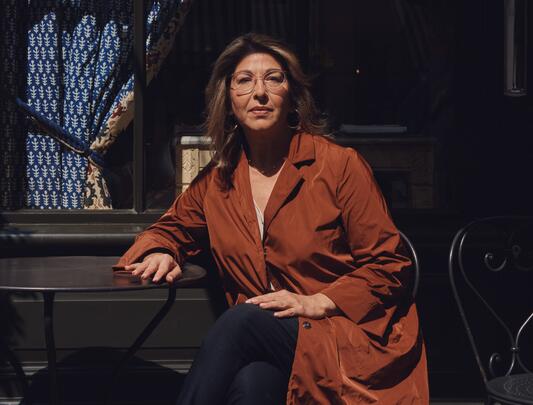 A photo of Naomi Klein seated at a metal table against a dark background, wearing glasses and a russet overcoat.
