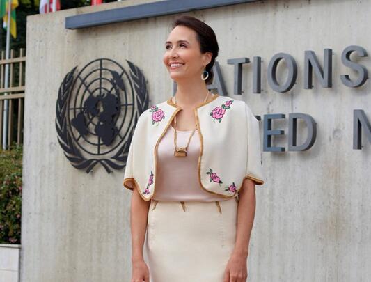 Standing in front of a United Nations sign, Jennifer Duncan wears a white blazer and skirt with rose designs and a gold trim.