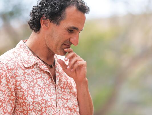 Simon Donner in a pink collared shirt, touching his chin with his finger, against a blurred grassy background.