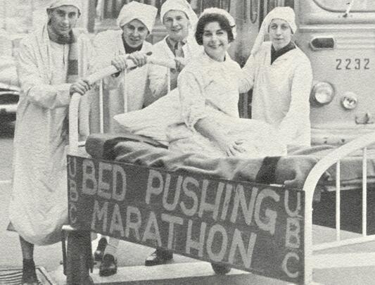  Four people standing around a person in a bed. Attached to the bed is a sign that reads: "UBC Bed Pushing Marathon"