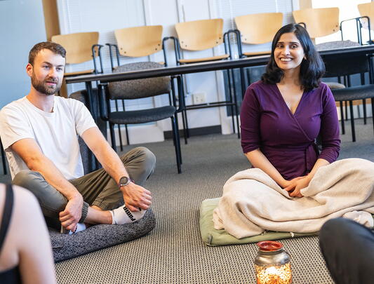 Dr. Varada Kolhatkar (right) seated cross-legged on a classroom floor, with other students seated in similar fashion