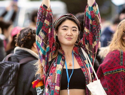 An image of Sophia Yang standing in front of a crowd with her arms up, wearing a lanyard, black top, and quilted red, green, and white top.