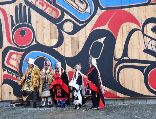 Five people in Indigenous outfits in front of wall with Indigenous artwork
