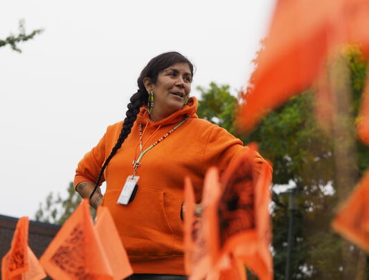 Indigenous woman dressed in orange stands behind small orange flags