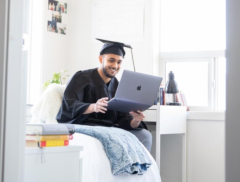 Student in cap and gown at home, looking at laptop