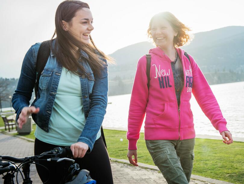 Two women walking along the lakefront. Woman on left has a bike with her.