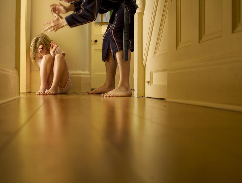 A child cowers in a corner while an adult's hands are positioned over her.