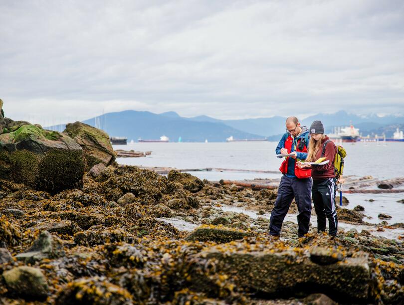 Two researchers by the water, jotting down notes, with a view of the mountains in the background.