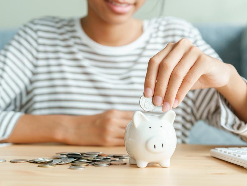 Piggy bank with coins. A person in the background is putting a coin in the piggy bank.