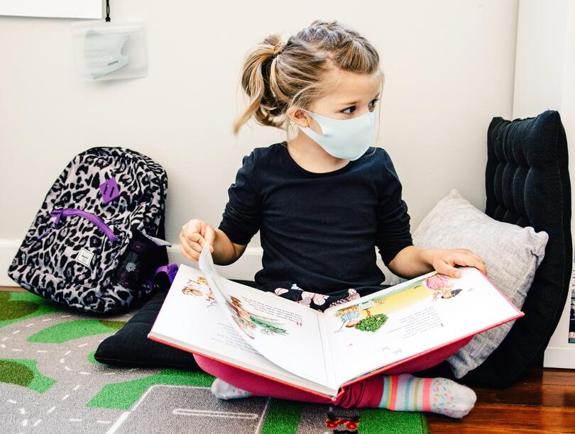 Photo of a masked child sitting on the ground reading.
