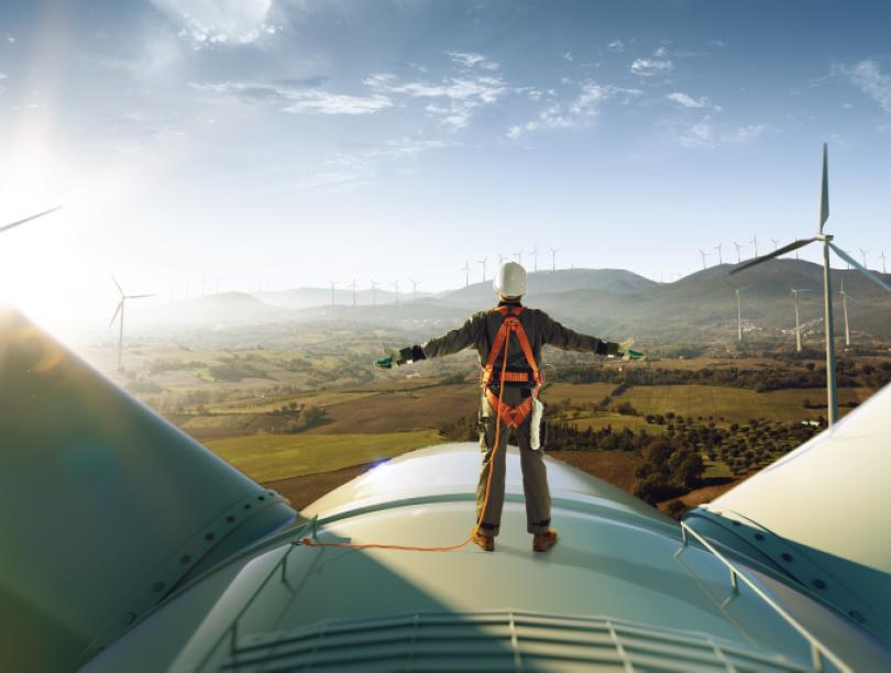 A person stands on a wind turbine, overlooking a wind farm. 
