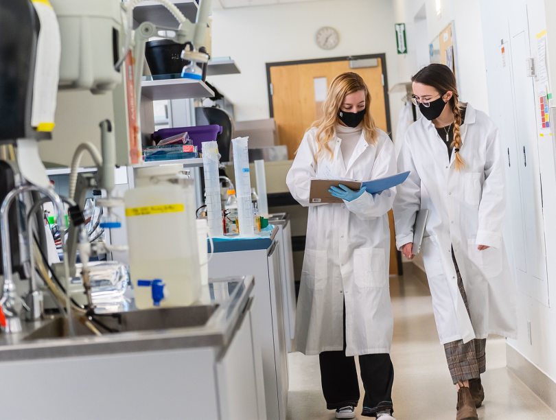 Two female students in labcoats walk in a laboratory.