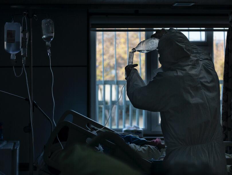 Nurse in PPE suit pours liquid into tube at patient's bed in hospital