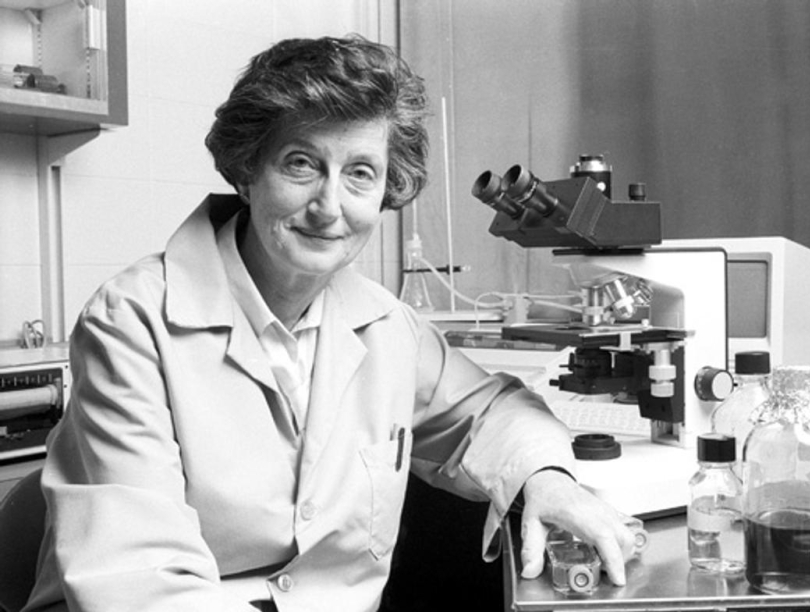 Black and white photo of Dr. Nelly Auersperg seated at a desk with microscope