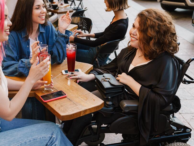 Three female friends with drinks around a table, including one in a wheelchair