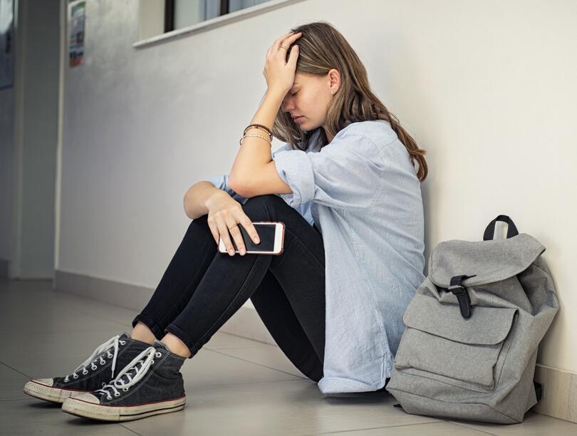 Young woman sitting against wall on hallway floor with hand on head and holding smartphone