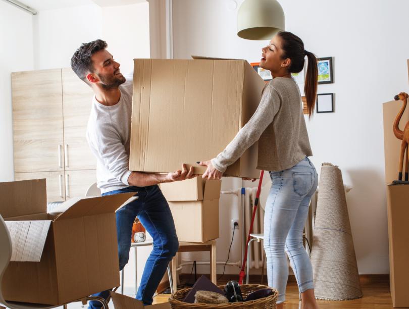 Man and woman carrying box through room amid other boxes and furniture